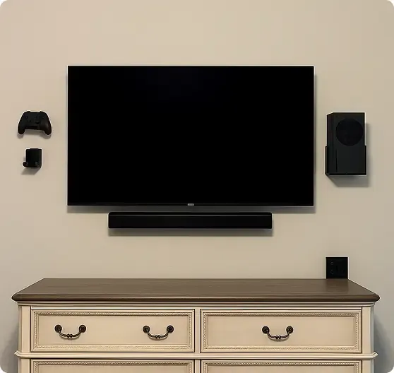 A wall-mounted flatscreen TV with a soundbar below, flanked by a game controller, remote, and speaker, above a cream-colored dresser with four drawers.