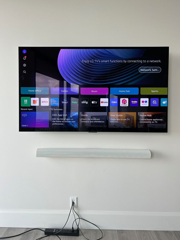 A wall-mounted LG smart TV displays its main home screen interface. Below is a white soundbar and a visible power cord plugged into an outlet.