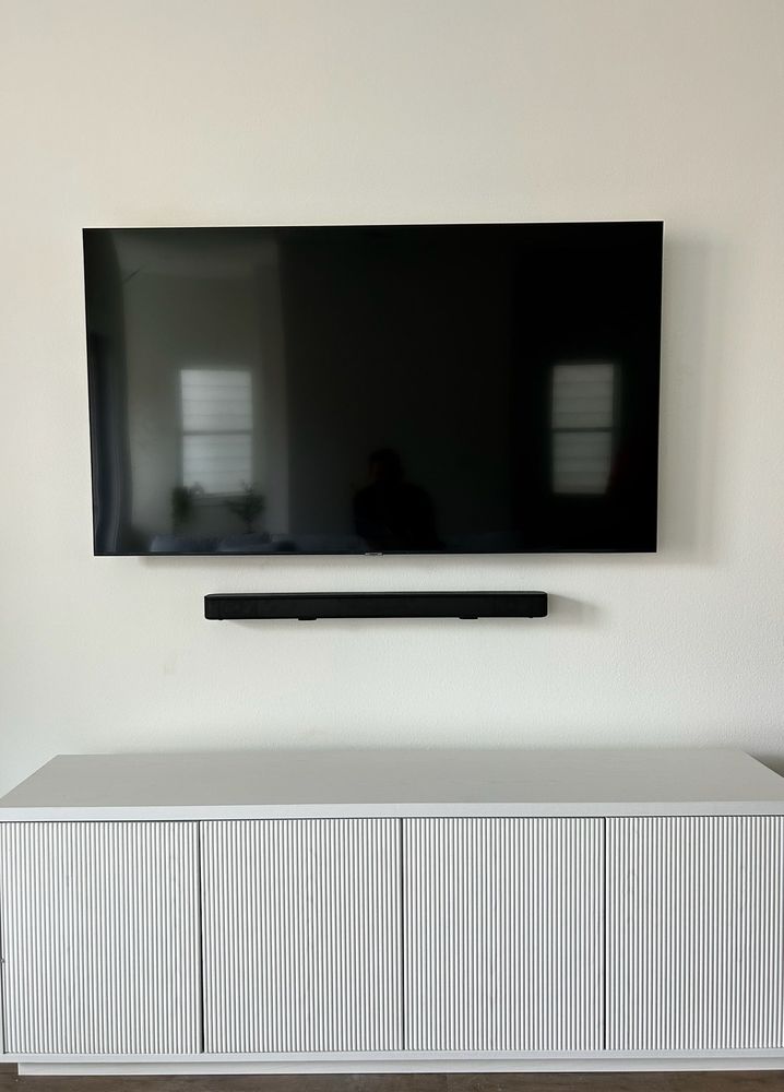 A flat-screen TV is mounted on a plain white wall above a black soundbar and a light-colored cabinet with vertical grooves.