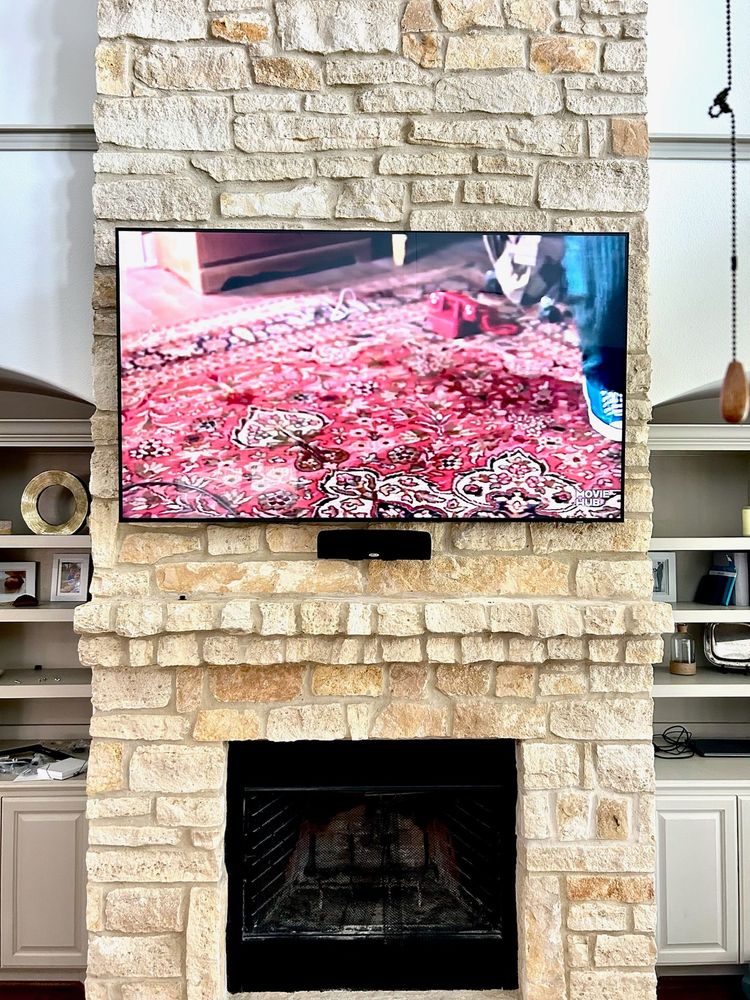 A flat-screen TV mounted on a stone fireplace shows a patterned rug. Built-in shelves with decor and cabinets flank the fireplace.