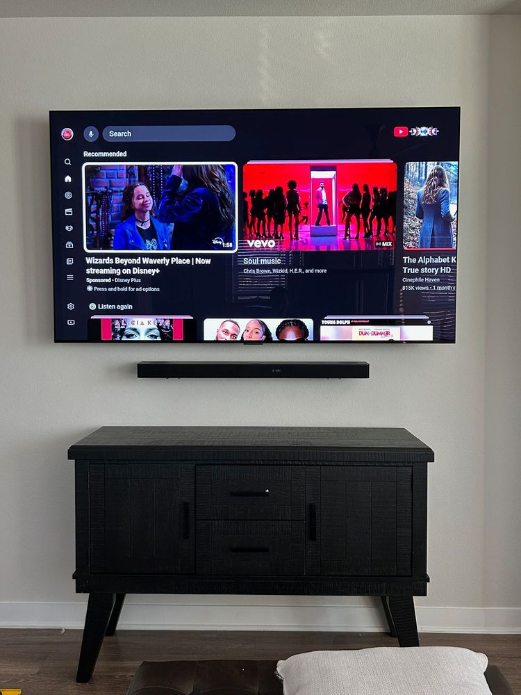 A flat-screen TV on a wall displays a streaming service interface above a black cabinet with a drawer and two doors.