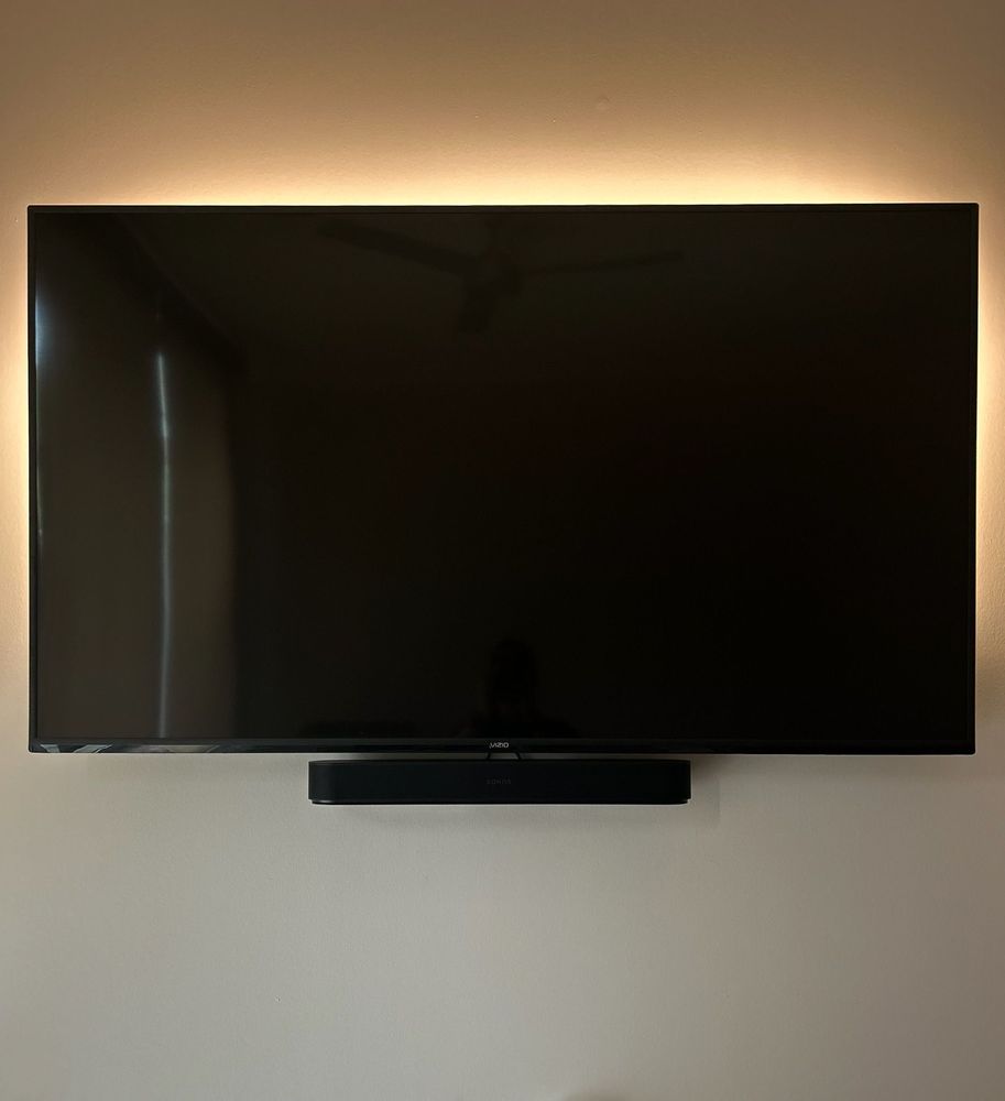 A large flat-screen TV is mounted on a beige wall above a black soundbar, with faint ambient lighting around the screen.
