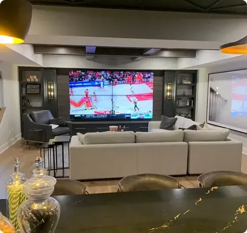 A modern living room with a large sectional sofa facing a big-screen TV showing a basketball game, with shelves, armchair, and countertop with glass jars in the foreground.