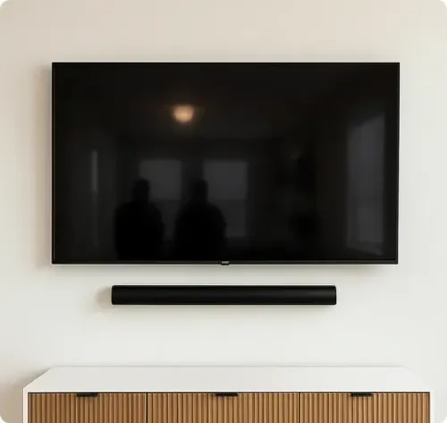 A flat-screen TV is mounted on a white wall above a soundbar, with a wooden cabinet below. Two faint reflections are visible on the TV screen.